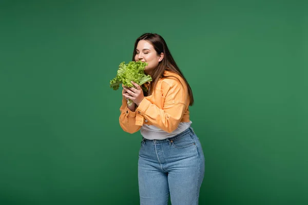 Sonriente mujer de tamaño grande ojos cerrados mientras sostiene la lechuga aislada en verde - foto de stock