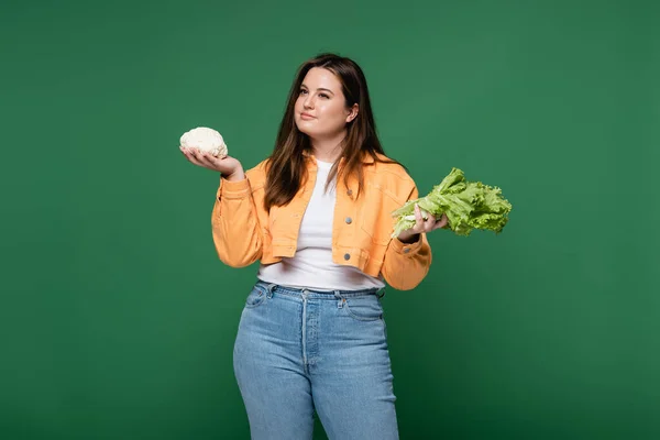 Mujer bonita con sobrepeso sosteniendo coliflor y lechuga aislada en verde - foto de stock