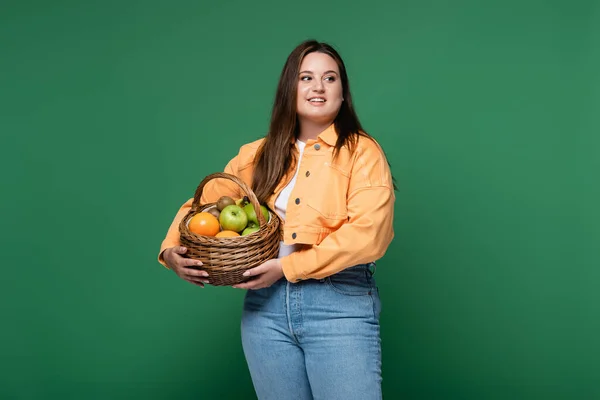 Mujer sonriente con cesta de sobrepeso con frutas aisladas en verde - foto de stock