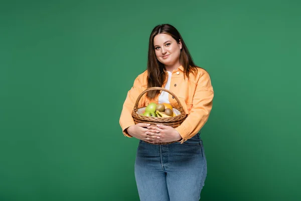 Молода жінка з надмірною вагою тримає кошик з фруктами і дивиться на камеру ізольовано на зеленому — стокове фото