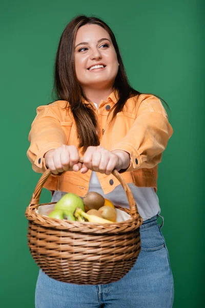 Mujer sonriente con sobrepeso sosteniendo canasta borrosa con frutas aisladas en verde - foto de stock