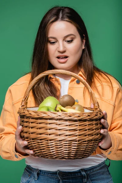 Astonished woman with overweight holding basket with fruits isolated on green — Stock Photo