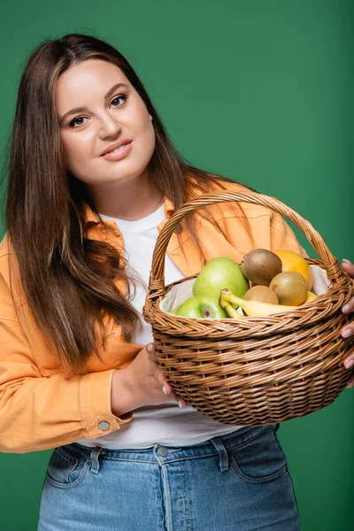 Mujer morena con cesta de sobrepeso con frutas aisladas en verde - foto de stock