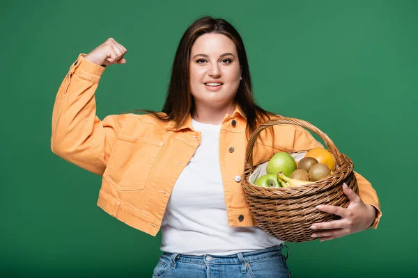 Mujer sonriente con sobrepeso mostrando músculos y sosteniendo cesta con frutas aisladas en verde - foto de stock