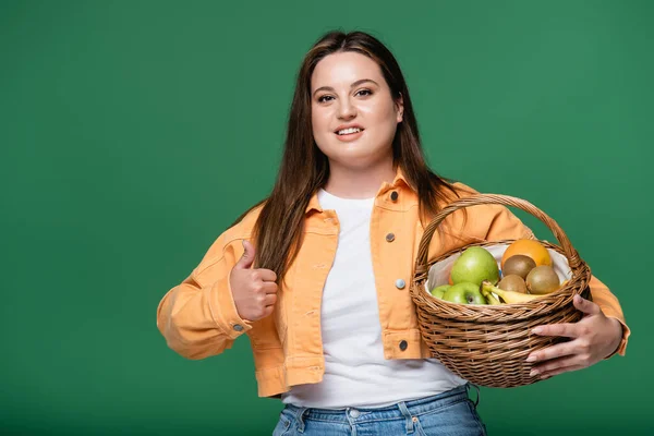 Mujer bonita con sobrepeso mostrando como y sosteniendo cesta con frutas aisladas en verde - foto de stock