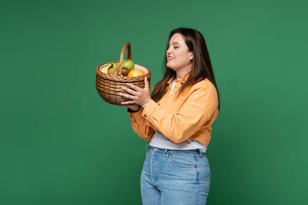 Mulher positiva com excesso de peso segurando cesta com frutas frescas isoladas em verde — Fotografia de Stock