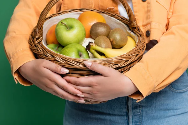 Обрезной вид женщины с лишним весом, держащей корзину со свежими фруктами, изолированными на зеленом — стоковое фото