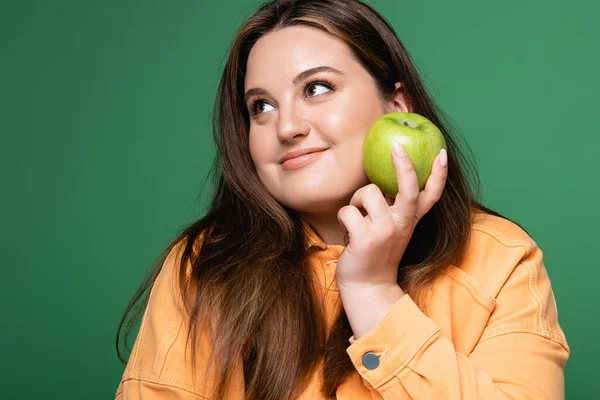 Веселая брюнетка с избыточным весом держит свежее яблоко изолированы на зеленый — стоковое фото