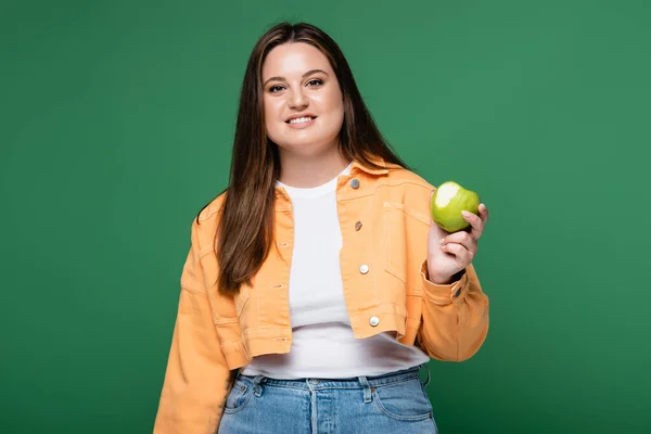 Mujer bonita cuerpo positivo sosteniendo manzana y sonriendo aislado en verde - foto de stock