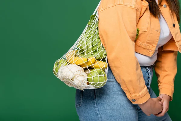 Обрезанный вид женщины с избыточным весом с сеткой мешок и свежие продукты питания изолированы на зеленый — стоковое фото