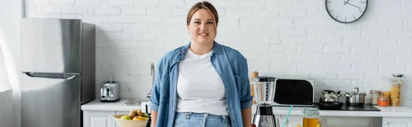 Позитивная женщина с избыточным весом стоит рядом с едой на кухне, баннер — стоковое фото