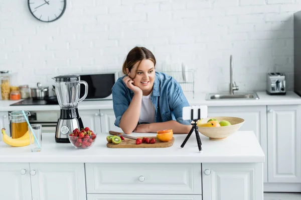 Mujer sonriente con sobrepeso mirando el teléfono inteligente cerca de frutas y licuadora en la mesa de la cocina - foto de stock