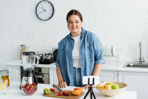 Donna con sovrappeso guardando smartphone vicino a frutta fresca e frullatore in cucina — Foto stock