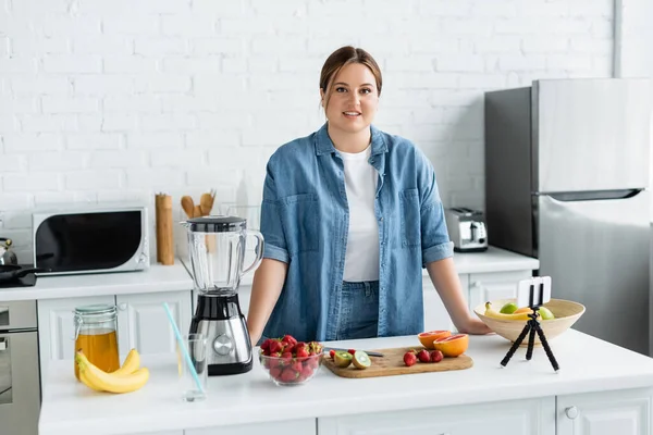 Mujer alegre cuerpo positivo mirando la cámara cerca de frutas, miel y teléfono inteligente en la cocina - foto de stock