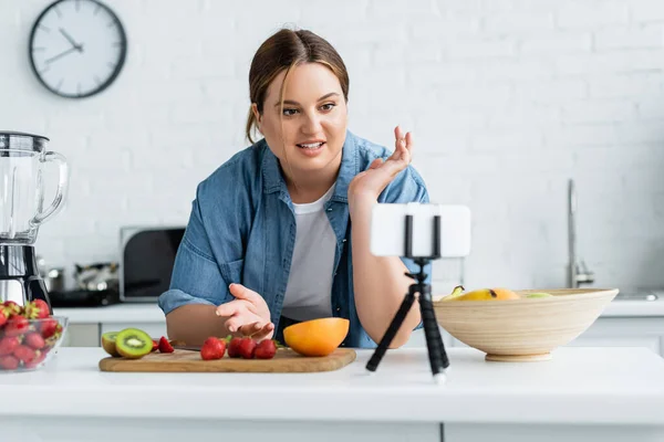 Lächelnder Blogger mit Übergewicht schaut in der Küche neben frischem Obst aufs Smartphone — Stockfoto