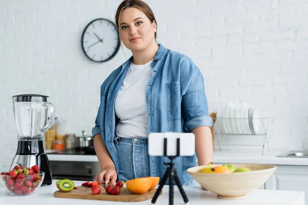Mujer de tamaño grande mirando la cámara cerca de un teléfono inteligente borroso y frutas maduras en la cocina - foto de stock