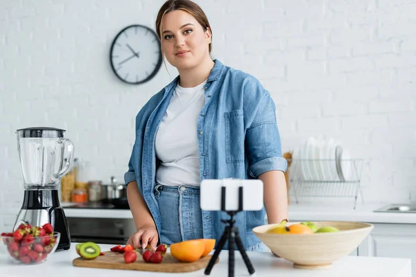 Plus size woman looking at camera near blurred smartphone and ripe fruits in kitchen — Stock Photo