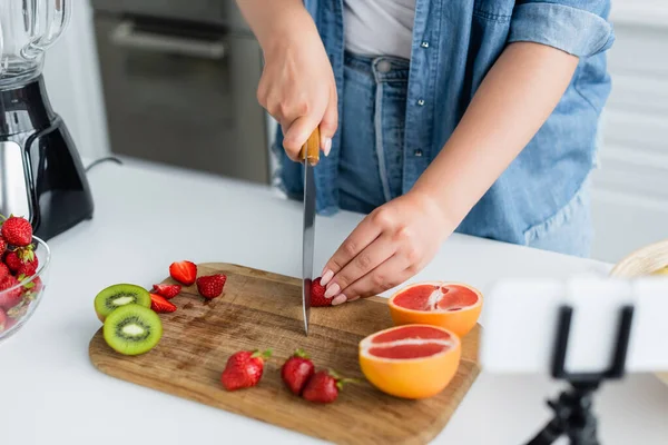 Vista recortada de la mujer joven cortando frutas frescas cerca de licuadora y teléfono inteligente borroso en la cocina - foto de stock