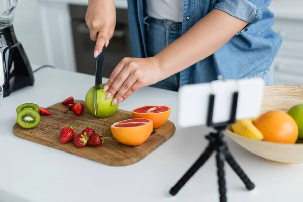 Vista ritagliata di giovane donna che taglia frutta vicino smartphone su treppiede in cucina — Foto stock