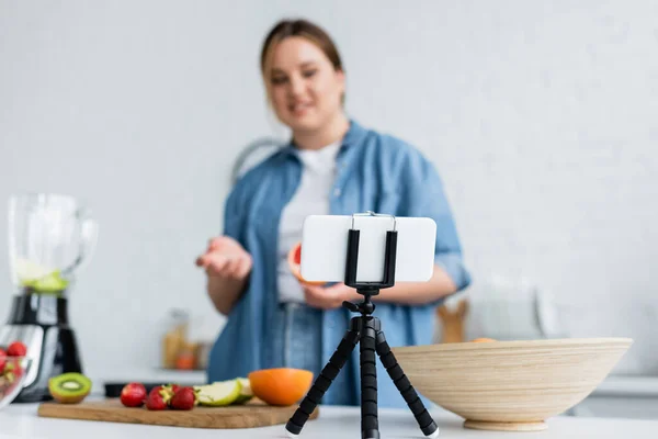 Smartphone en trípode cerca borrosa más tamaño mujer y frutas en la cocina - foto de stock