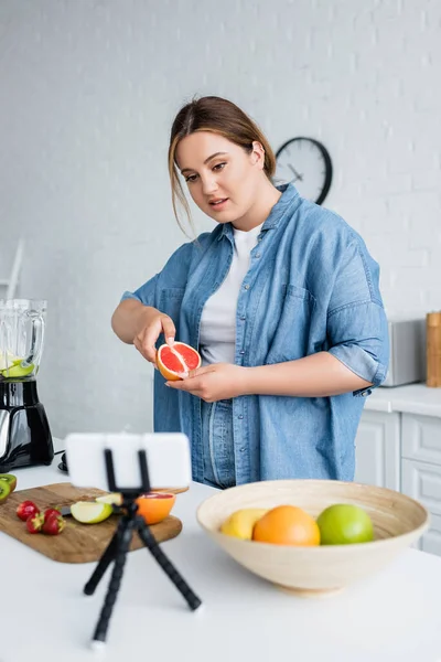 Grande taille femme tenant pamplemousse près flou smartphone et mélangeur dans la cuisine — Photo de stock