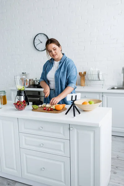 Sonriente mujer de tamaño grande cortando frutas frescas cerca de licuadora y teléfono inteligente en la cocina - foto de stock