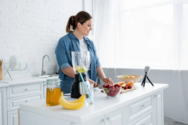 Vista lateral de mujer de talla grande cortando frutas maduras cerca de smartphone y licuadora en casa - foto de stock