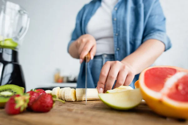 Vista recortada de mujer borrosa con sobrepeso corte plátano cerca de frutas y licuadora en la cocina — Stock Photo