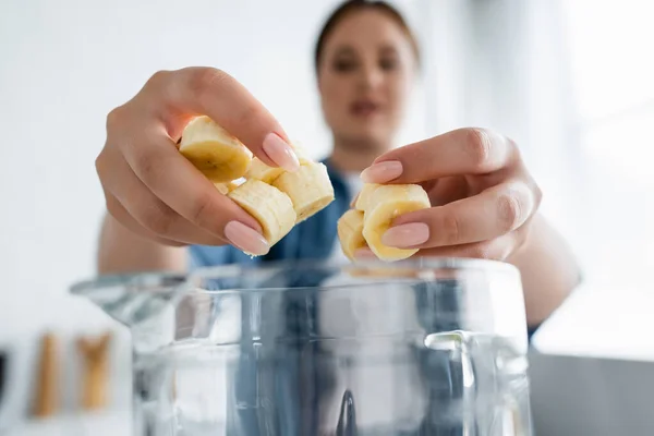 Donna offuscata che tiene la banana affettata vicino al frullatore in cucina — Foto stock