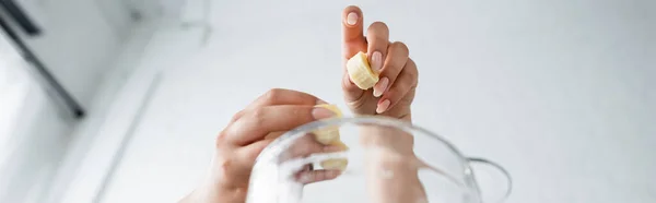 Vue du bas de la femme tenant une banane tranchée près du mélangeur dans la cuisine, bannière — Photo de stock