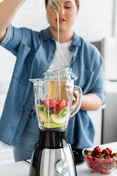 Blurred plus size mulher derramando mel em frutas no liquidificador na cozinha — Fotografia de Stock