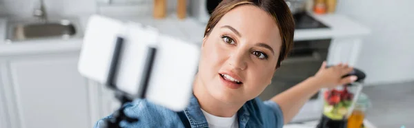 Vista de ángulo alto de la mujer sonriente de tamaño grande usando un teléfono inteligente borroso cerca de la licuadora borrosa, pancarta - foto de stock