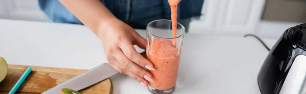 Cropped view of blurred woman pouring smoothie in glass in kitchen, banner — Stock Photo