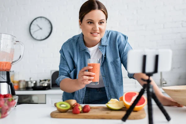 Positivo más tamaño mujer sosteniendo vidrio de batido cerca de teléfono celular borroso en trípode en la cocina - foto de stock