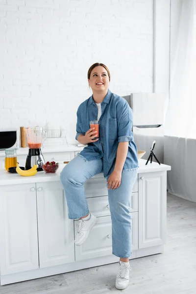 Feliz mujer de tamaño grande sosteniendo batido cerca de frutas, licuadora y teléfono inteligente en la cocina - foto de stock