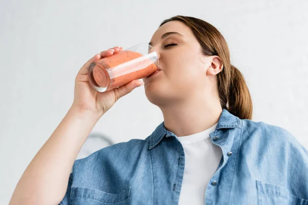Низкий угол обзора молодой женщины с избыточным весом питьевого смузи дома — стоковое фото
