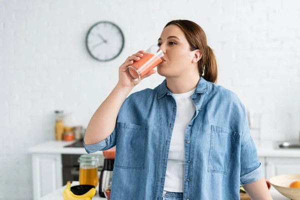 Plus size woman drinking fruit smoothie in kitchen — Stock Photo