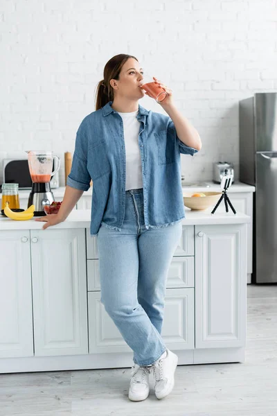 Giovane donna con frullato da bere in sovrappeso vicino al cellulare e frutta matura in cucina — Foto stock