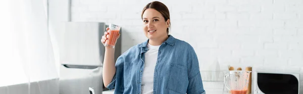 Happy plus taille femme avec verre de smoothie regardant la caméra près du mélangeur dans la cuisine, bannière — Photo de stock