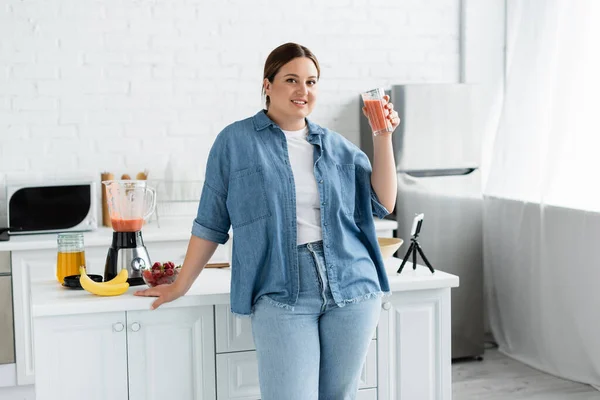 Улыбающаяся женщина plus size держит стакан смузи и смотрит на камеру возле мобильного телефона на кухне — стоковое фото