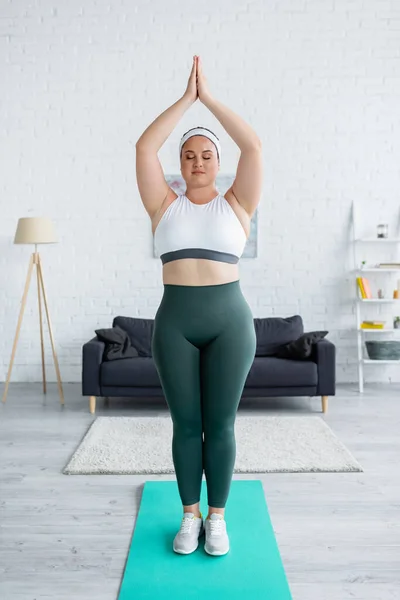 Plus size woman in sportswear standing with praying hands on fitness mat — Stock Photo