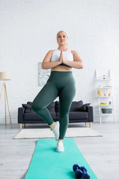 Grande taille femme debout avec des mains priantes sur tapis de fitness près des haltères à la maison — Photo de stock