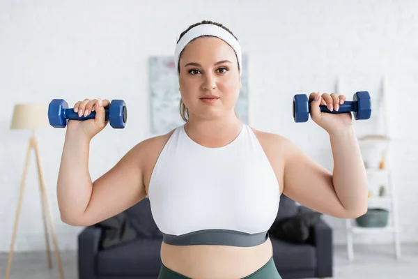 Молодая женщина с избыточным весом упражнения с гантелями и смотреть в камеру дома — стоковое фото