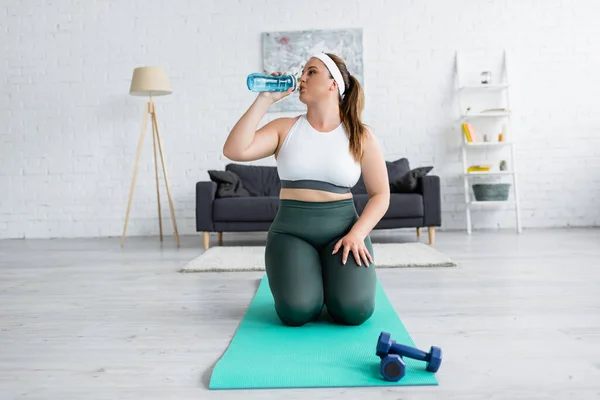 Plus Size Frau in Sportbekleidung trinkt Wasser in der Nähe von Fitnessmatte und Kurzhanteln zu Hause — Stockfoto