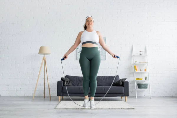 Sorrindo mais tamanho mulher treinamento com pular corda na sala de estar — Fotografia de Stock