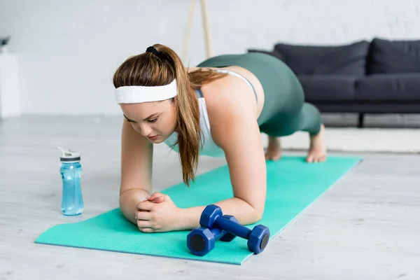 Plus-Size-Frau steht in Planke auf Fitnessmatte neben Hanteln und Sportflasche zu Hause — Stockfoto