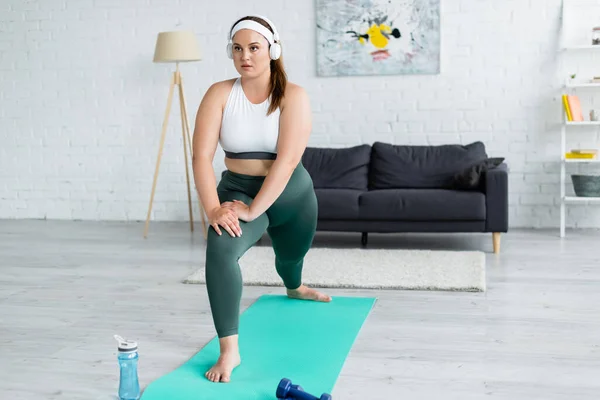 Giovane sportiva plus size in cuffia che si esercita sul tappeto fitness vicino alla bottiglia sportiva in soggiorno — Foto stock