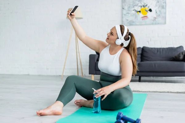 Вид сбоку улыбающейся plus size женщины в наушниках, делающей селфи на смартфоне рядом со спортивной бутылкой на фитнес-коврике — стоковое фото