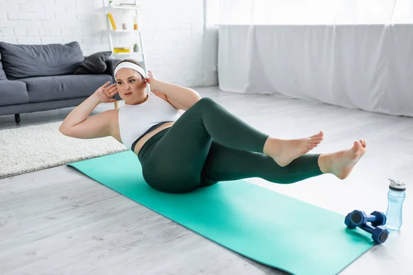 Jeune sportive pieds nus avec entraînement en surpoids près des haltères et bouteille de sport dans le salon — Photo de stock