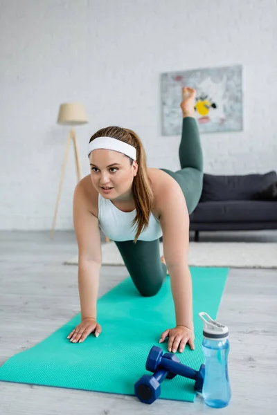 Sonriente deportista de talla grande haciendo ejercicio cerca de pesas y botellas deportivas en casa - foto de stock